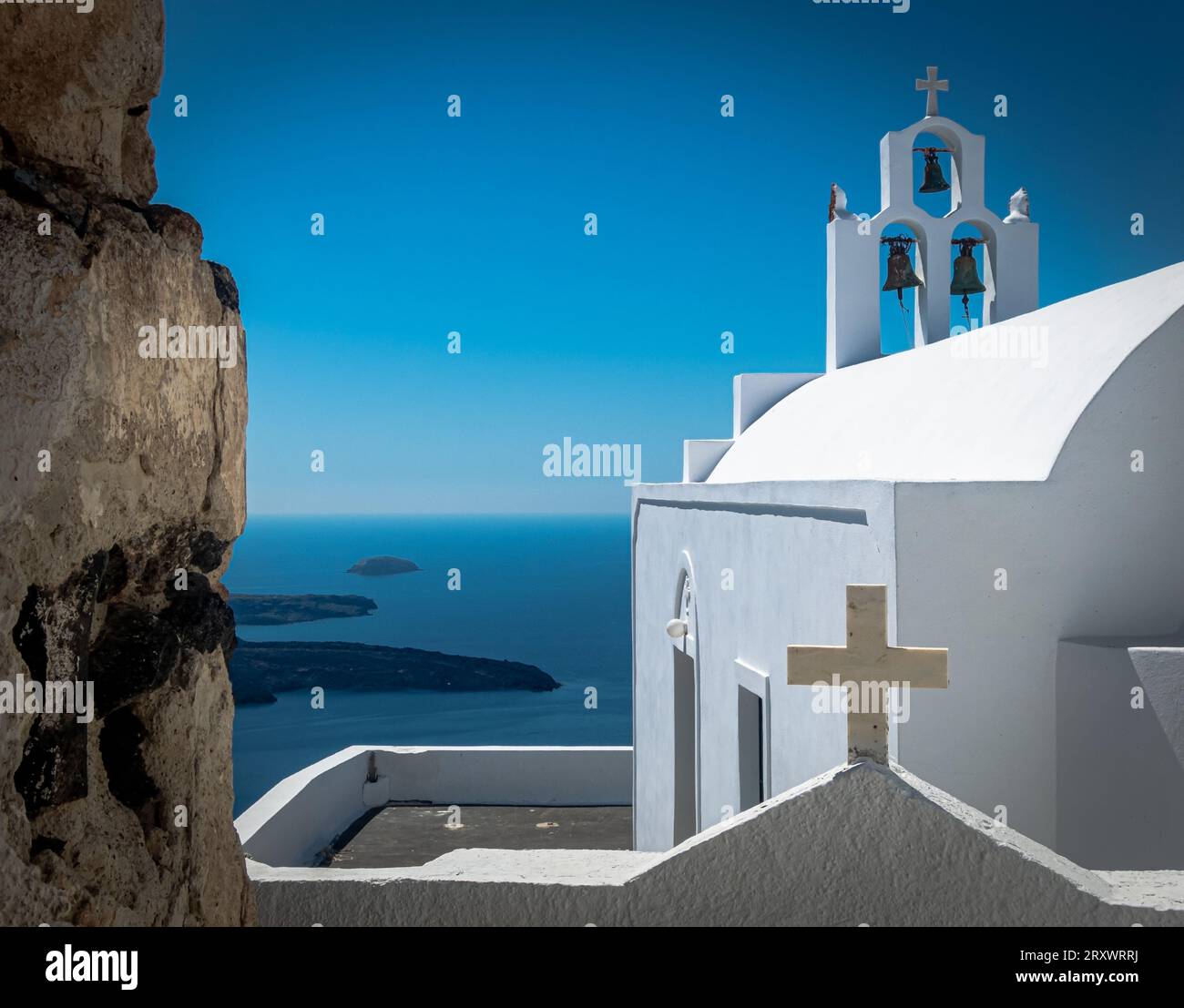 La chiesa bianca di Santorini si affaccia sul mare con 3 campane in un piccolo campanile e croci religiose. Niente persone. Posizione alta e cielo limpido Foto Stock