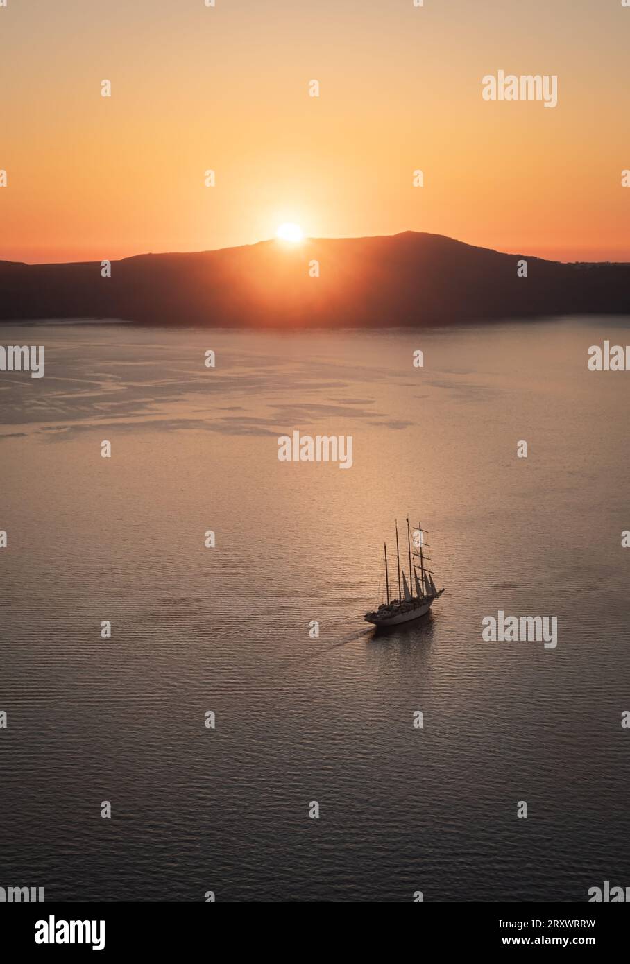 La baia al largo di Thera, Santorini, con il sole che tramonta dietro l'isola di Thirasia, il cielo limpido e dorato e la barca a vela alberata nel mezzo della baia Foto Stock