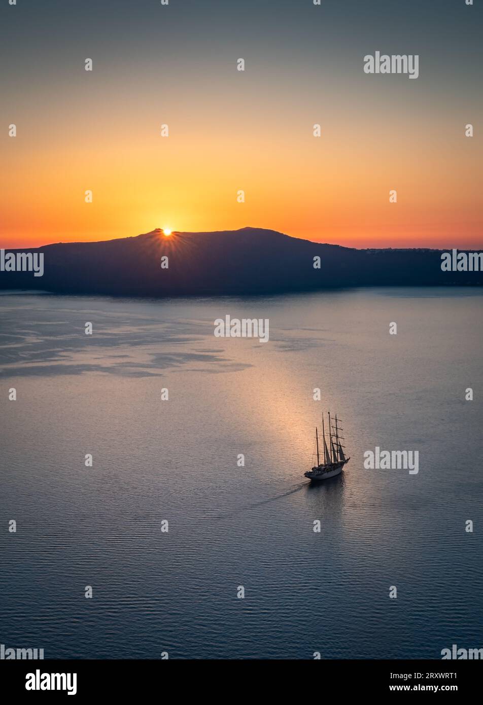 La baia al largo di Thera, Santorini, con il sole che tramonta dietro l'isola di Thirasia, il cielo limpido e dorato e la barca a vela alberata nel mezzo della baia Foto Stock