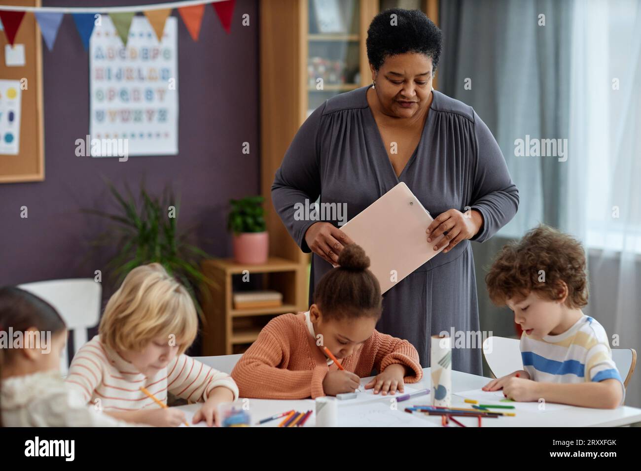 Ritratto di gentile donna nera che guarda bambini piccoli mentre fanno arte e artigianato in classe prescolare, spazio per le copie Foto Stock