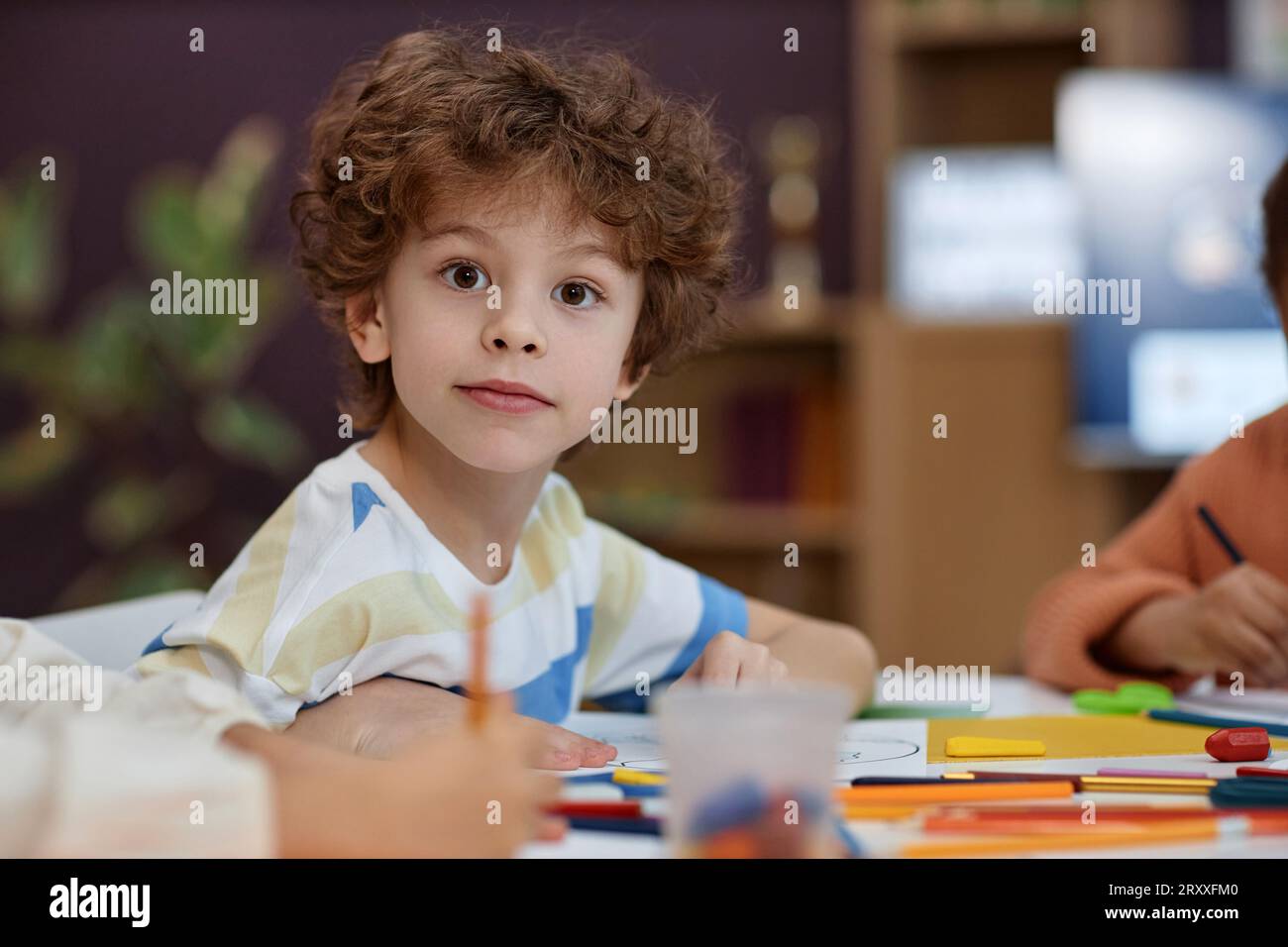 Ritratto di un bambino carino che guarda la macchina fotografica mentre colora le foto al tavolo durante la lezione di arte e artigianato all'asilo, nello spazio per le copie Foto Stock