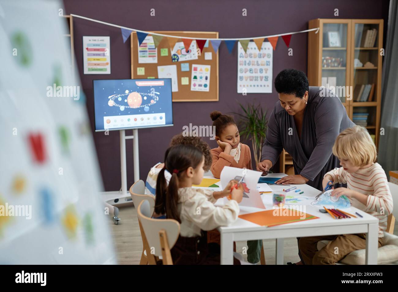 Ritratto di gentile donna nera che aiuta un gruppo di bambini seduti a un tavolo in classe prescolare, spazio copia Foto Stock