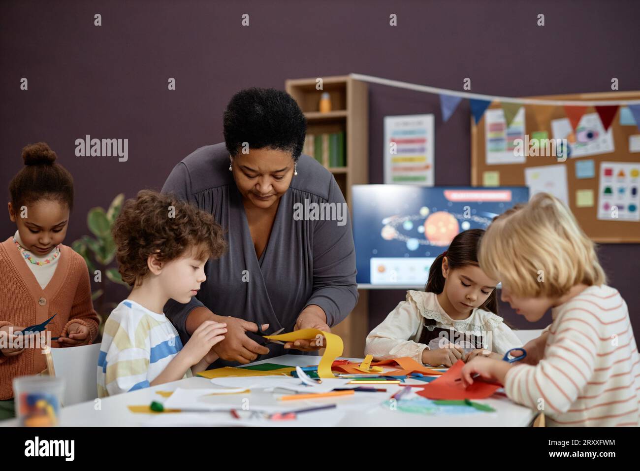 Ritratto di gentile donna nera che aiuta un gruppo di bambini piccoli a fare artigianato in classe prescolare, spazio di copia Foto Stock