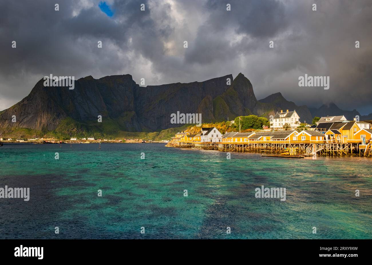 Pittoresco villaggio di Lofoten con tradizionali palafitte e una catena montuosa sullo sfondo. La scena è immersa nella luce del sole ed è accompagnata Foto Stock
