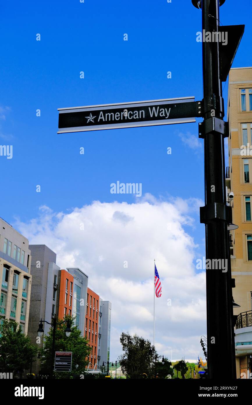 Cartello stradale "American Way" contro un cielo blu con nuvole bianche. Una bandiera americana è appesa sullo sfondo. Foto Stock