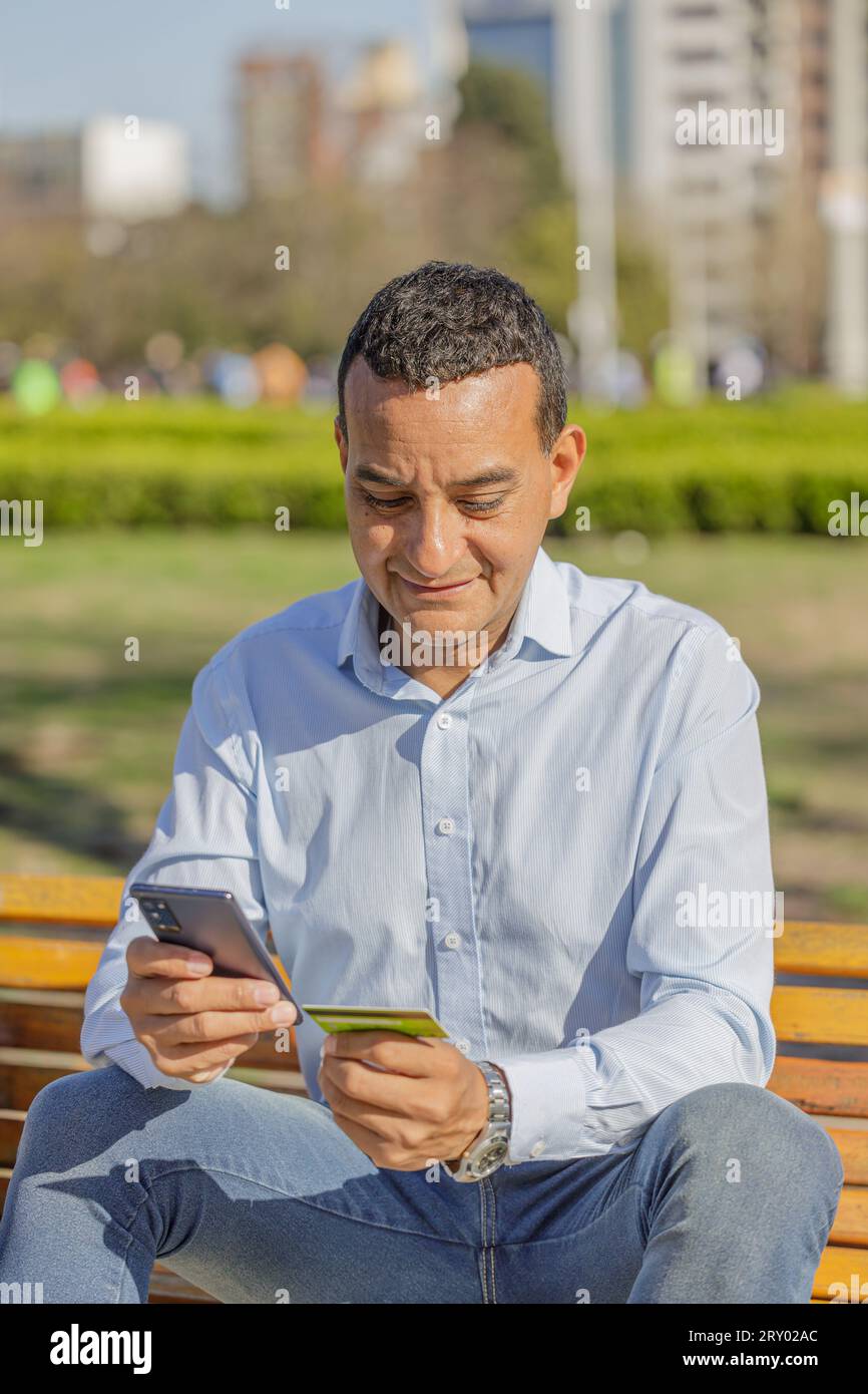 Un giovane latinoamericano che fa un acquisto con una carta di credito sul suo cellulare in un parco pubblico. Foto Stock