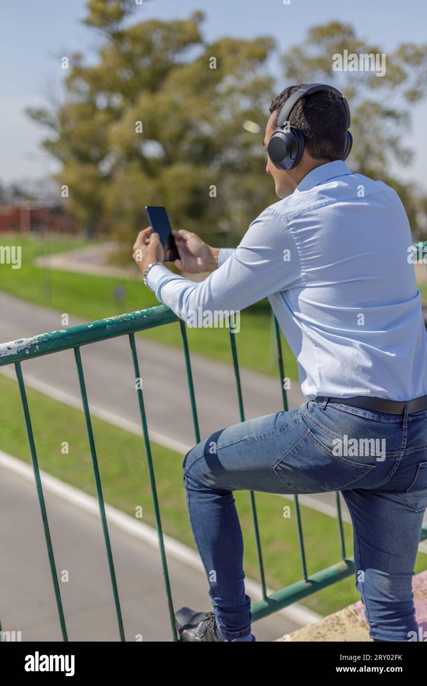 Uomo latino con le cuffie che usa il suo telefono cellulare appoggiato alla ringhiera di un ponte pedonale. Foto Stock