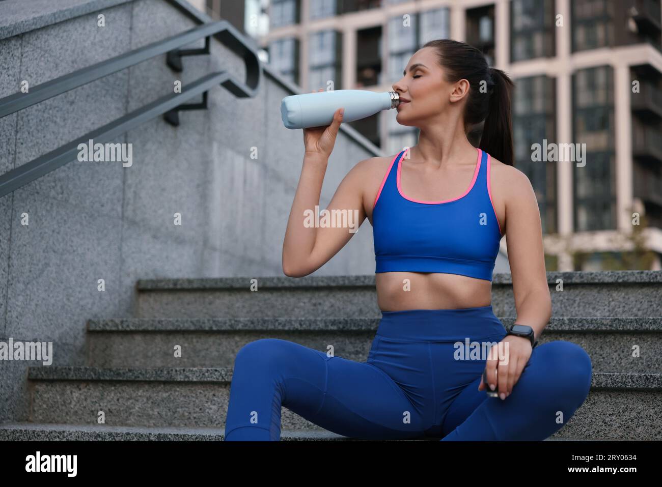 Bella donna con elegante abbigliamento sportivo, acqua potabile sulle scale all'aperto Foto Stock