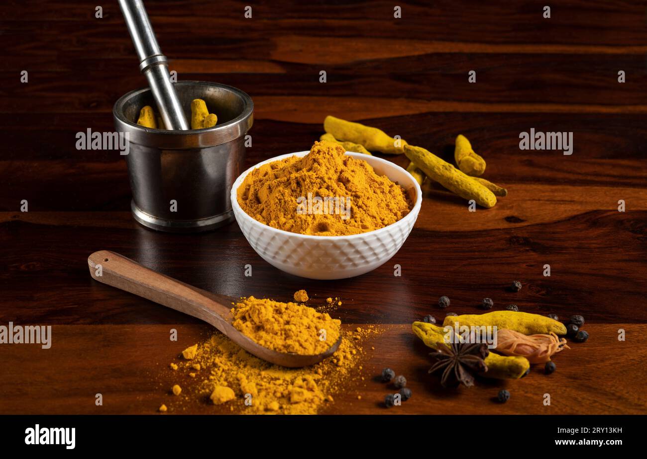 Polvere di curcuma conservata in una ciotola bianca e cucchiaio di legno con Malta e pestello, radice secca di curcuma, spezie secche tenute in giro come sostegno su una surfa di legno marrone Foto Stock