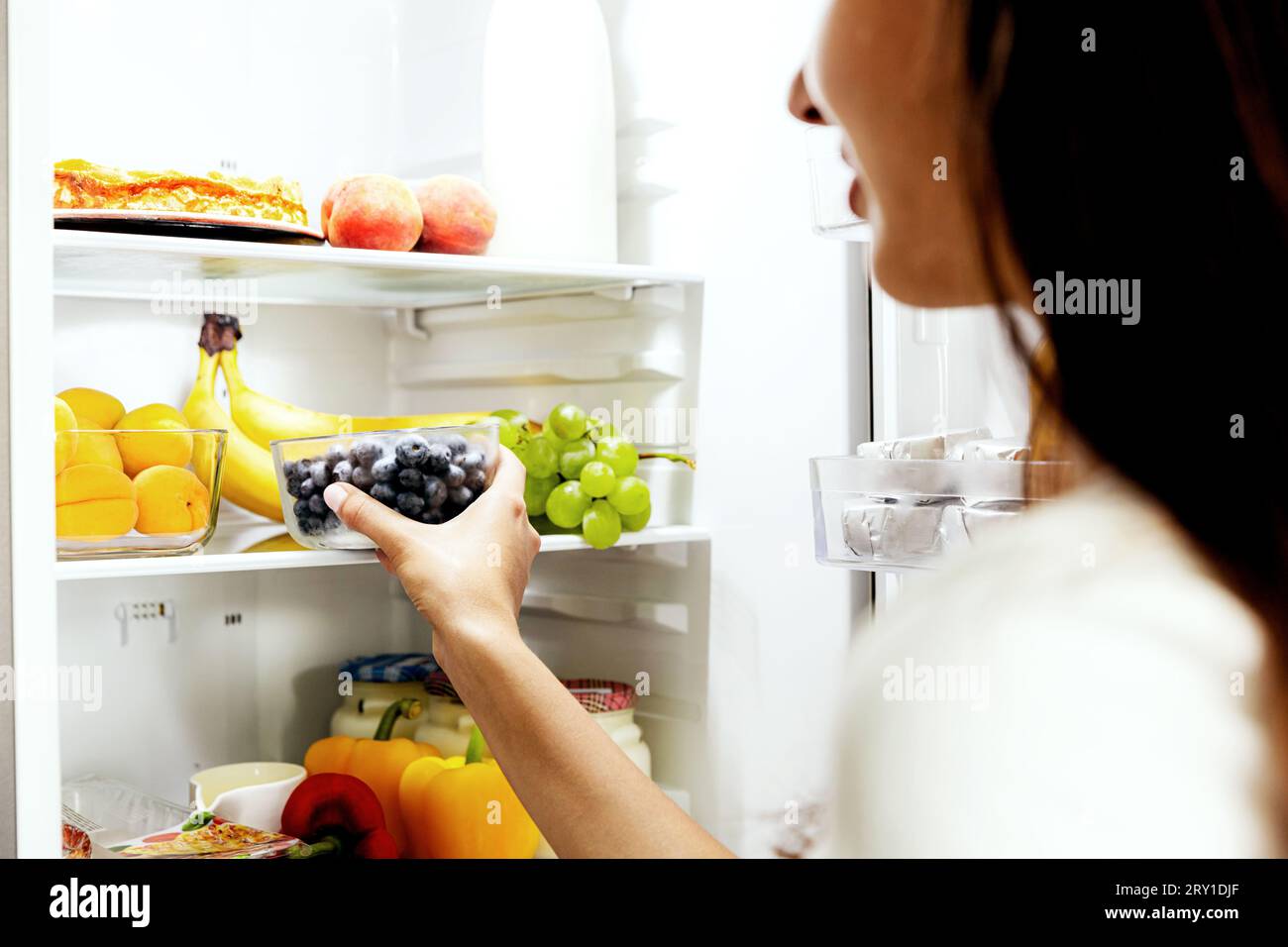 Donna che prende, afferra o preleva una scatola di mirtilli dal ripiano aperto del frigorifero o dal cassetto del frigorifero pieno di frutta, verdura, banana, pesche, Foto Stock