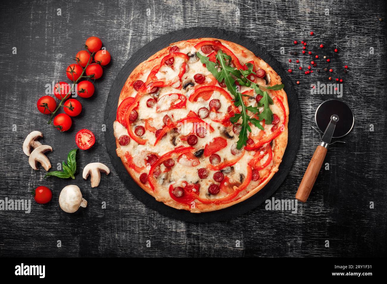 Pizza con salsicce affumicate, formaggio, funghi, pomodori ciliegini, peperoni e verdure su una pietra e un gesso nero, vista dall'alto. Foto Stock