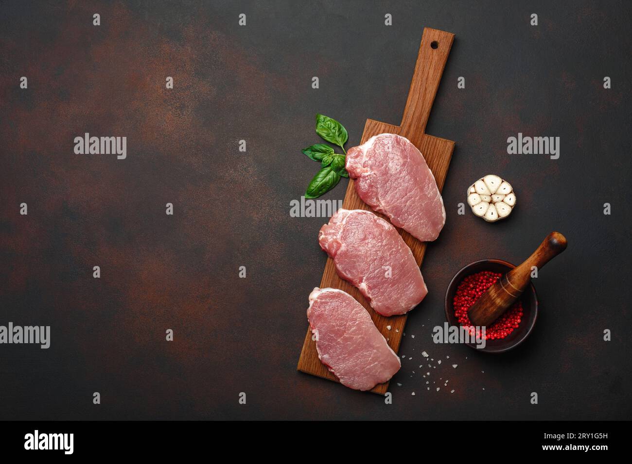 Pezzi di carne di maiale cruda con basilico, aglio, pepe, sale e mortaio di spezie su tagliere e fondo marrone arrugginito. Vista dall'alto con spazio per la tua t Foto Stock