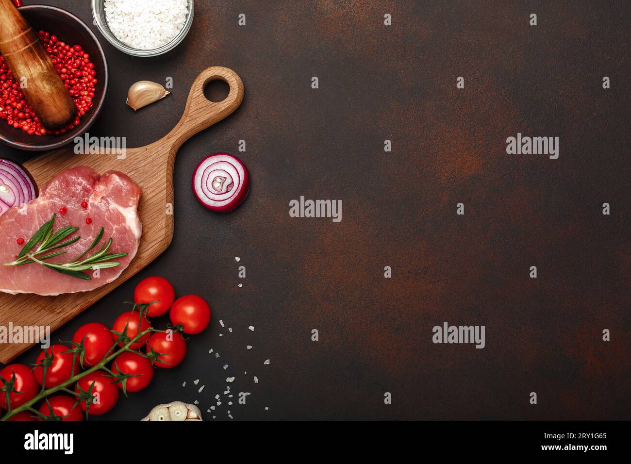 Pezzi di bistecca di maiale cruda su tagliere con pomodori ciliegini, rosmarino, aglio, pepe rosso, cipolla, mortaio salino e spezie su fondo marrone arrugginito. Foto Stock