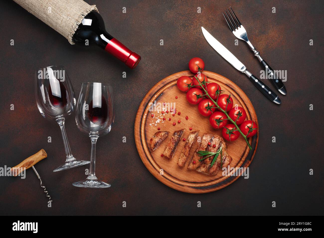 Bistecche di maiale grigliate a fette con bottiglia di vino, bicchiere di vino, cavatappi, coltello, forchetta, pane nero, pomodori ciliegini, aglio, cipolla e rosmarino su arrugginito Foto Stock