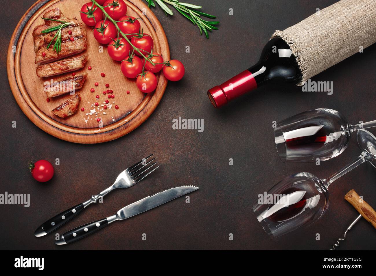 Bistecche di maiale grigliate a fette con bottiglia di vino, bicchiere di vino, cavatappi, coltello, forchetta, pane nero, pomodori ciliegini e rosmarino su fondo arrugginito. In alto Foto Stock