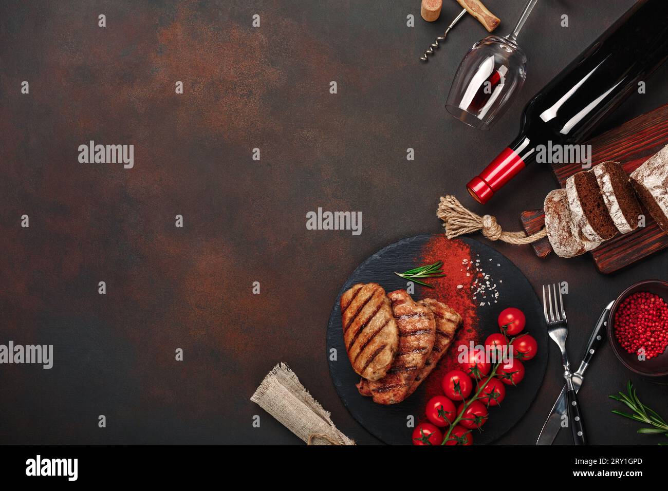 Bistecche di maiale alla griglia con bottiglia di vino, bicchiere di vino, coltello, forchetta, pane nero, pomodori ciliegini e rosmarino su fondo arrugginito. Vista dall'alto. Foto Stock