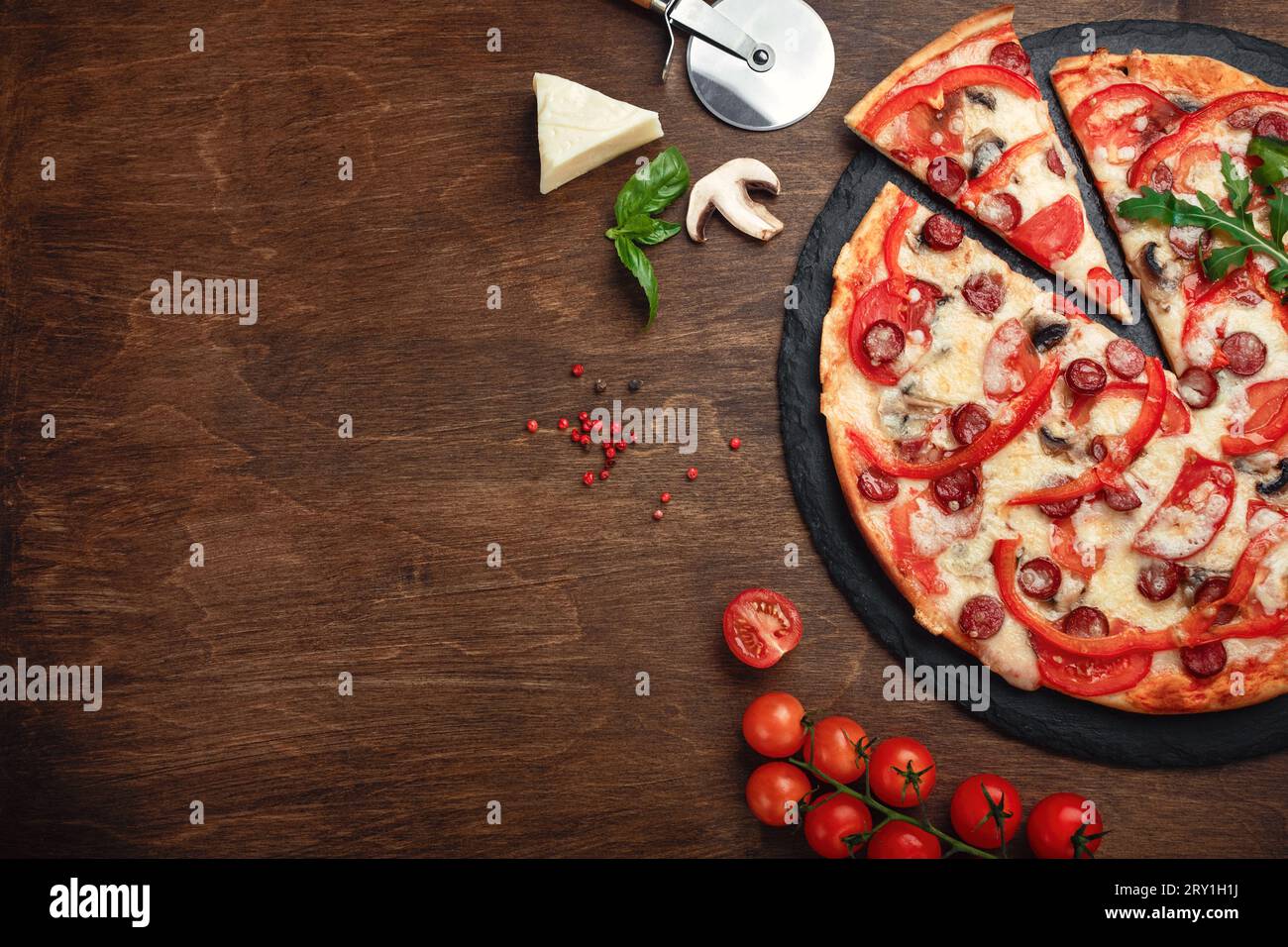 Pizza con salsicce affumicate, formaggio, funghi, pomodori ciliegini, peperoni e verdure su una pietra e una tavola marrone di legno, vista dall'alto. Foto Stock