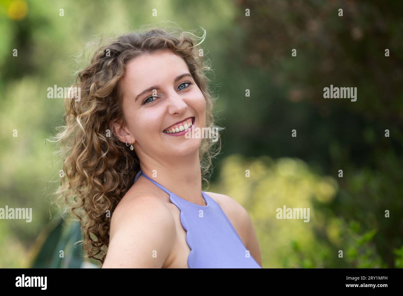 Ritratto della testa e delle spalle di una giovane donna caucasica sorridente con capelli biondi ricci e occhi blu, all'aperto in estate Foto Stock