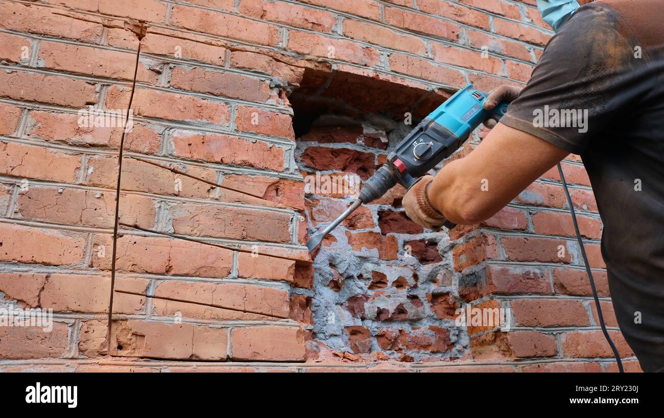 Un costruttore con una T-shirt nera e guanti che scalpellano la muratura di una casa con un trapano pneumatico, lavorando per distruggere un muro di mattoni Foto Stock