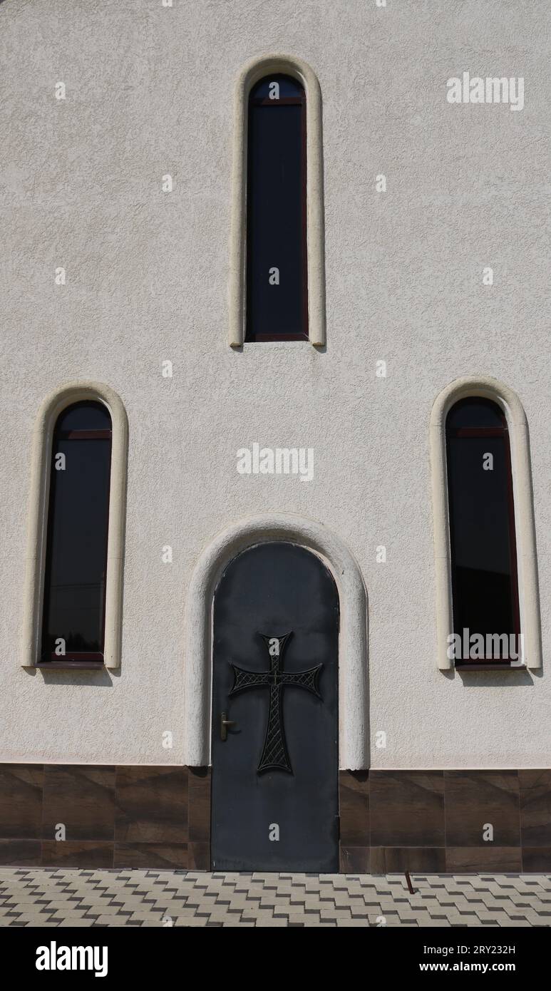 Parete chiara di un edificio religioso con finestre strette ad arco e una vecchia porta con croce cristiana, immagine verticale, frammento dell'esterno Foto Stock
