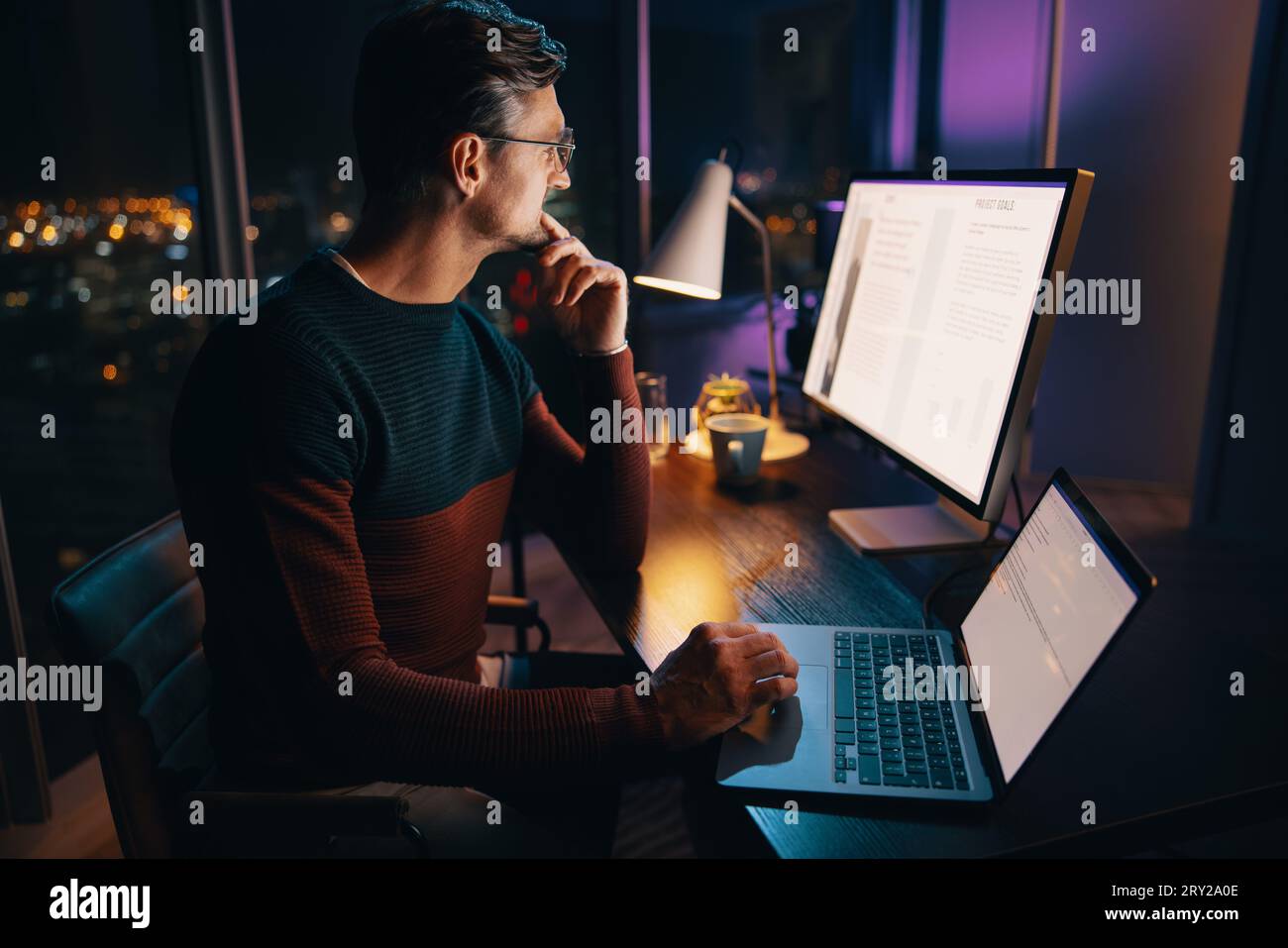 Un uomo d'affari caucasico lavora fino a tardi in ufficio, pianificando e pianificando un progetto di marketing sul suo portatile. L'atmosfera è tranquilla e premurosa Foto Stock