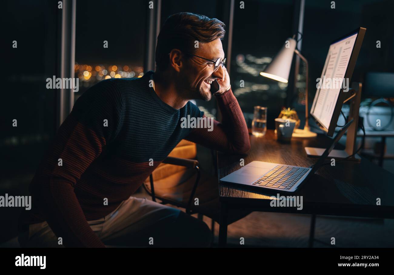 Un uomo d'affari felice lavora a tarda notte in ufficio, discutendo di un progetto durante una telefonata. Si siede alla sua scrivania, concentrato sul suo portatile, maintaini Foto Stock