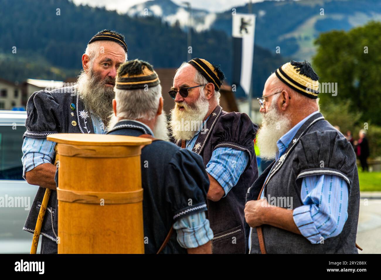 Un Barbu de la Gruyère porta un leggero recipiente di legno a forma di botte sulla schiena, probabilmente un "oji", un contenitore tradizionale usato nel dialetto della Gruyère per trasportare formaggio, mentre parla con tre uomini barbuti in costume durante l’Alpabzug a Oberschrot, Plaffeien, cantone di Friburgo, Svizzera. Tutti indossano la giacca ricamata Bredzon e il caratteristico berretto a strisce dei Barbus de la Gruyère. Foto Stock