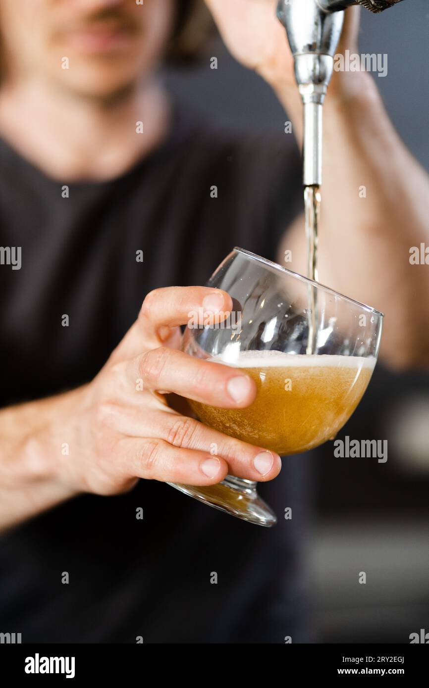 Anonimo giovane maschio non rasato che guarda in basso mentre si sta in piedi con il bicchiere trasparente aprendo il rubinetto e versando birra alla luce del giorno contro l'inte sfocato Foto Stock