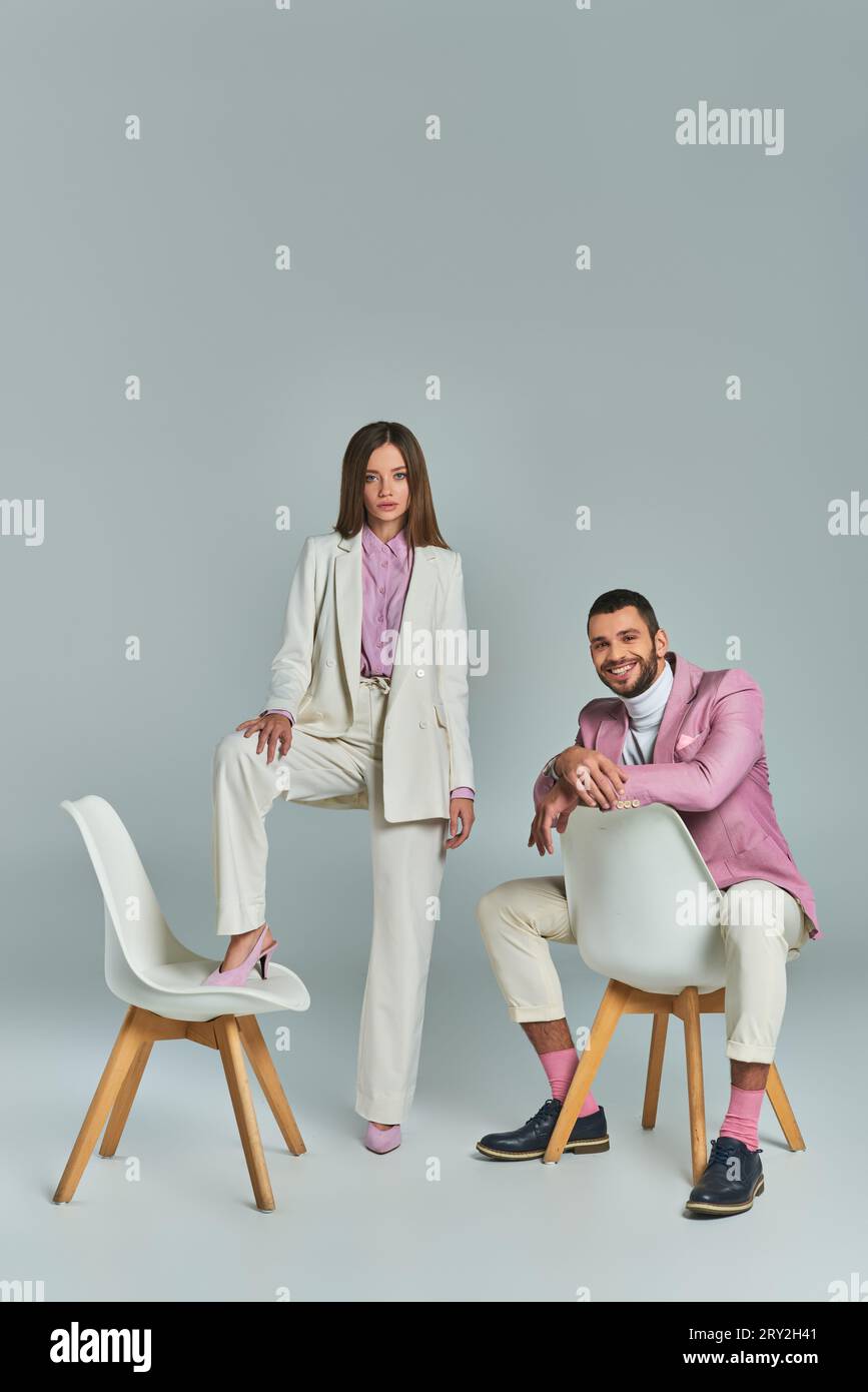 uomo gioioso con blazer lilla seduto e guardando la fotocamera vicino a una donna che posa con la poltrona grigia Foto Stock