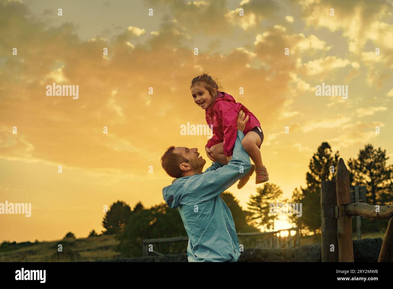 Al tramonto in una zona boscosa, un padre solleva con gioia la figlia di 3 anni, ridendo e crogiolandosi nella retroilluminazione dorata. Una recinzione rustica incornicia il Foto Stock