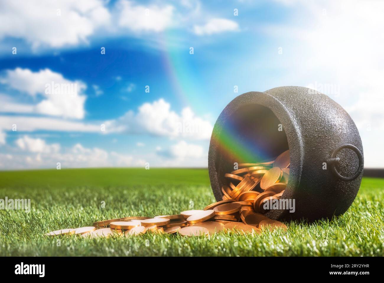 Angolo basso di un bollitore in ferro battuto nero che è stato rovesciato versando le sue monete d'oro sull'erba, con luce solare e arcobaleno. Foto Stock