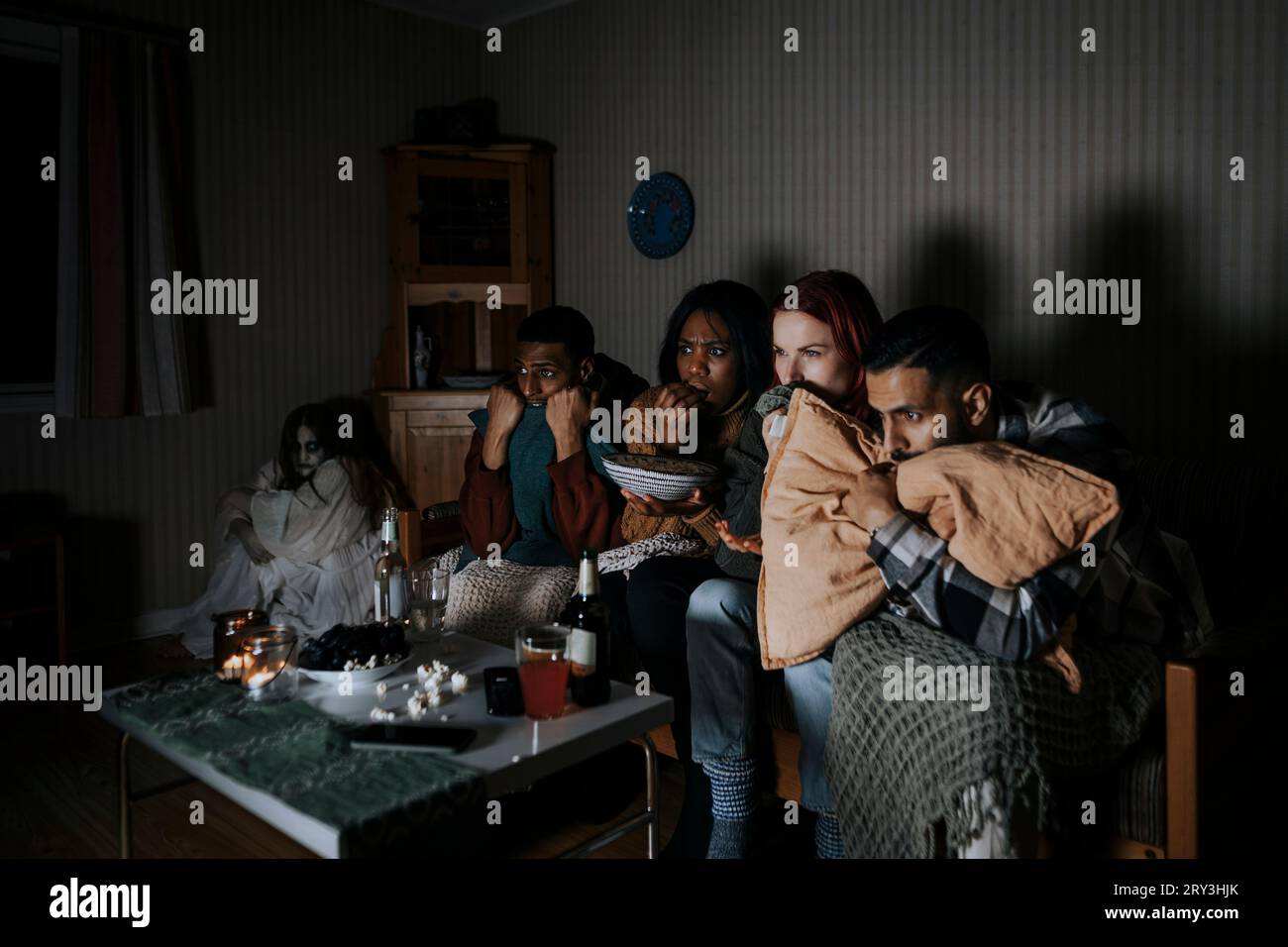 Amici multirazziali spaventati che guardano un film spaventoso mentre i fantasmi sono seduti in un angolo in cabina Foto Stock