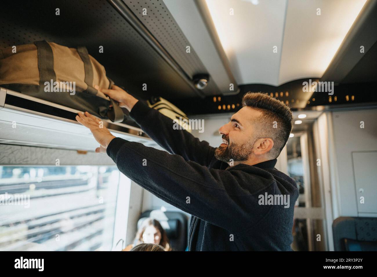 Vista laterale dell'uomo sorridente che carica i bagagli sullo scaffale del treno Foto Stock