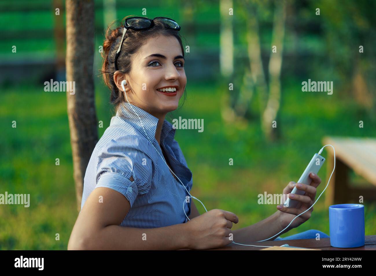 All'aperto, giovane donna lavora dinamicamente, gestendo telefono e laptop. In camicetta a righe, i suoi capelli sono tenuti da occhiali, in mezzo al paesaggio montano. Il suo expressi Foto Stock