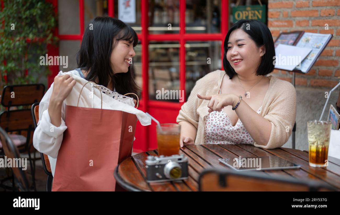 Una giovane e allegra donna asiatica si diverte a parlare con la sua amica mentre si rilassa a un tavolo all'aperto in una caffetteria durante la sua giornata di shopping Foto Stock