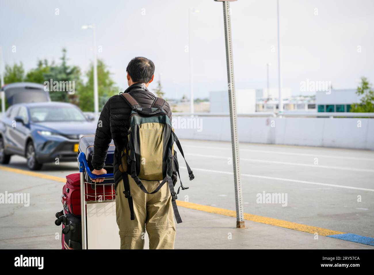 Uomo che spinge il carrello per i bagagli e cammina verso un'auto parcheggiata fuori dall'aeroporto. Foto Stock