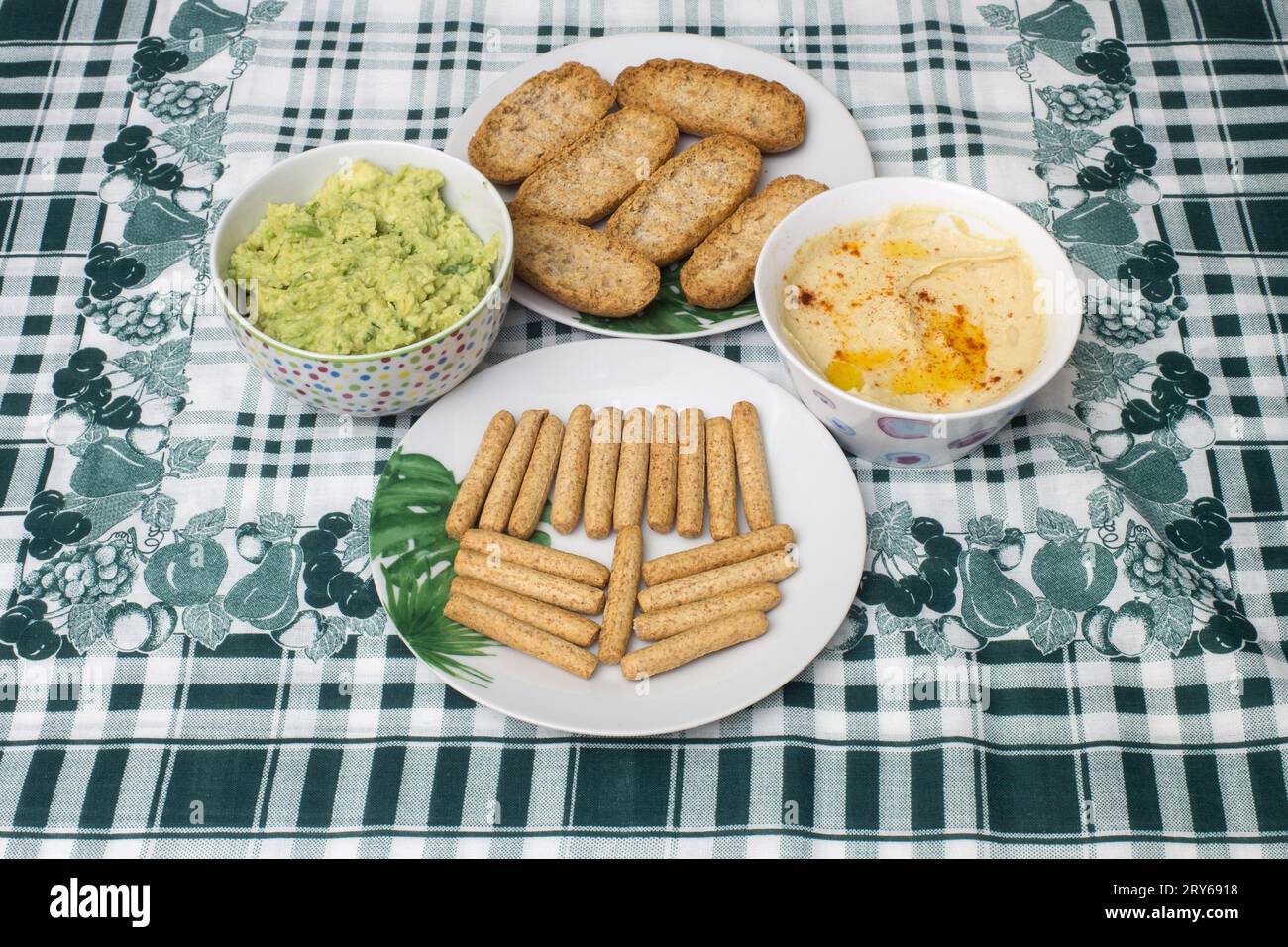 Due ciotole di hummus cremoso e salsa guacamole sono poste su un tavolo con una tovaglia a foglia e geometrica, insieme a toast di grano intero e grissini Foto Stock