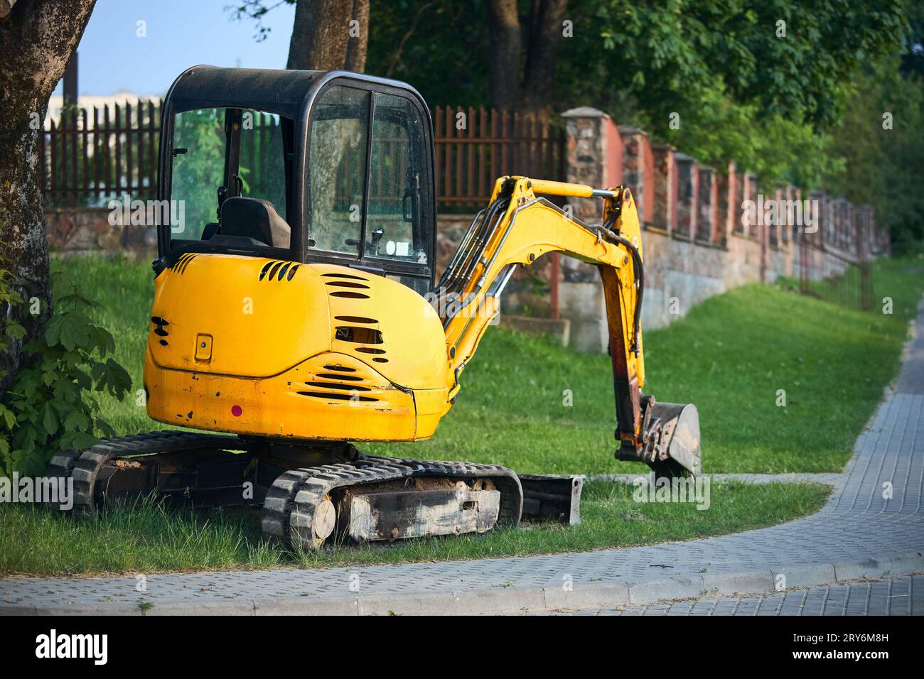 Scavatrice per miniescavatori arancione cingolata vicino alla strada con alberi verdi e recinzione in pietra sullo sfondo con spazio coperto. Foto Stock