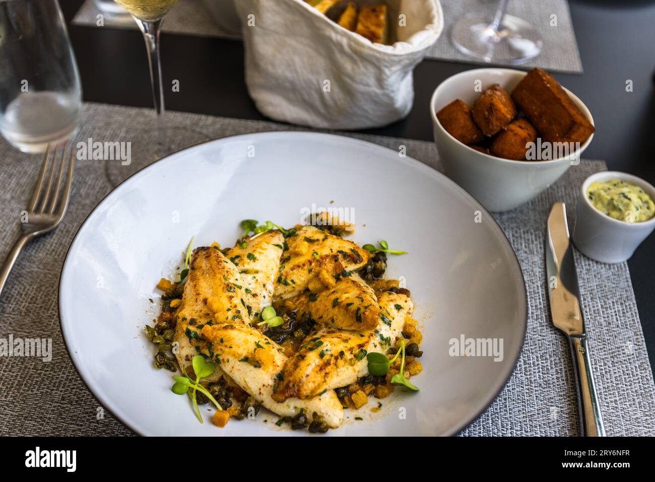Filetti di persico („Egli“) fritti si adagiano su cubetti dorati e capperi, guarniti con germogli di erbe, allo Château Salavaux a Vully-les-Lacs, nel cantone di Vaud, Svizzera – il piatto è legato al buono „Filetto di Egli con insalata“ del programma di viaggio „Genussreise – Étape Gourmande“. Accompagnano il piatto fette di patate dolci arrostite, burro alle erbe, pane in un canovaccio e un bicchiere di vino bianco. Il persico è considerato un pesce tipico dei laghi svizzeri. Foto Stock