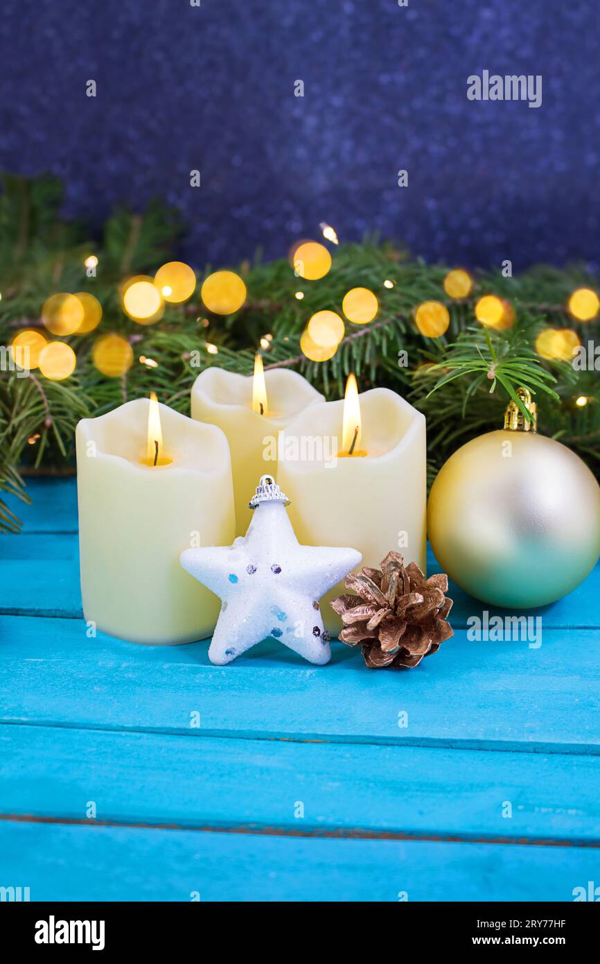 Candele di Natale con fuoco, stella bianca scintillante, cono di pino dorato e palla su sfondo di legno blu. Dietro ci sono rami di abete rosso con sfocatura Foto Stock
