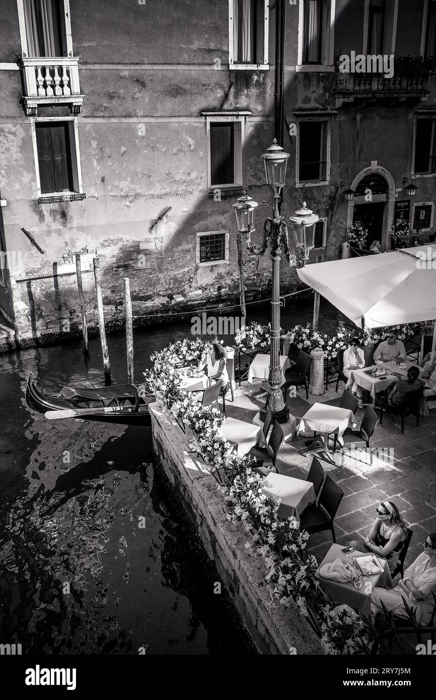 Vista in bianco e nero di una romantica caffetteria accanto a un canale veneziano con passaggio in gondola, catturando il fascino e l'eleganza senza tempo di Venezia, Italia Foto Stock