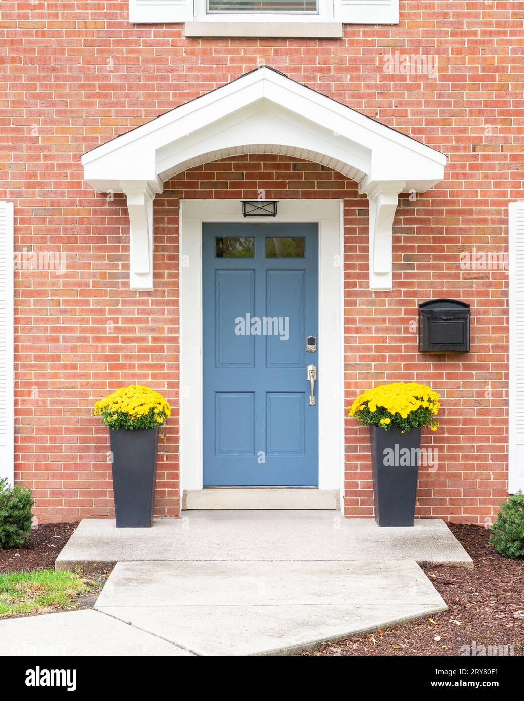 Una porta d'ingresso di una casa in mattoni rossi con uno sbalzo bianco, una porta d'ingresso blu e fioriere con fiori gialli. Foto Stock