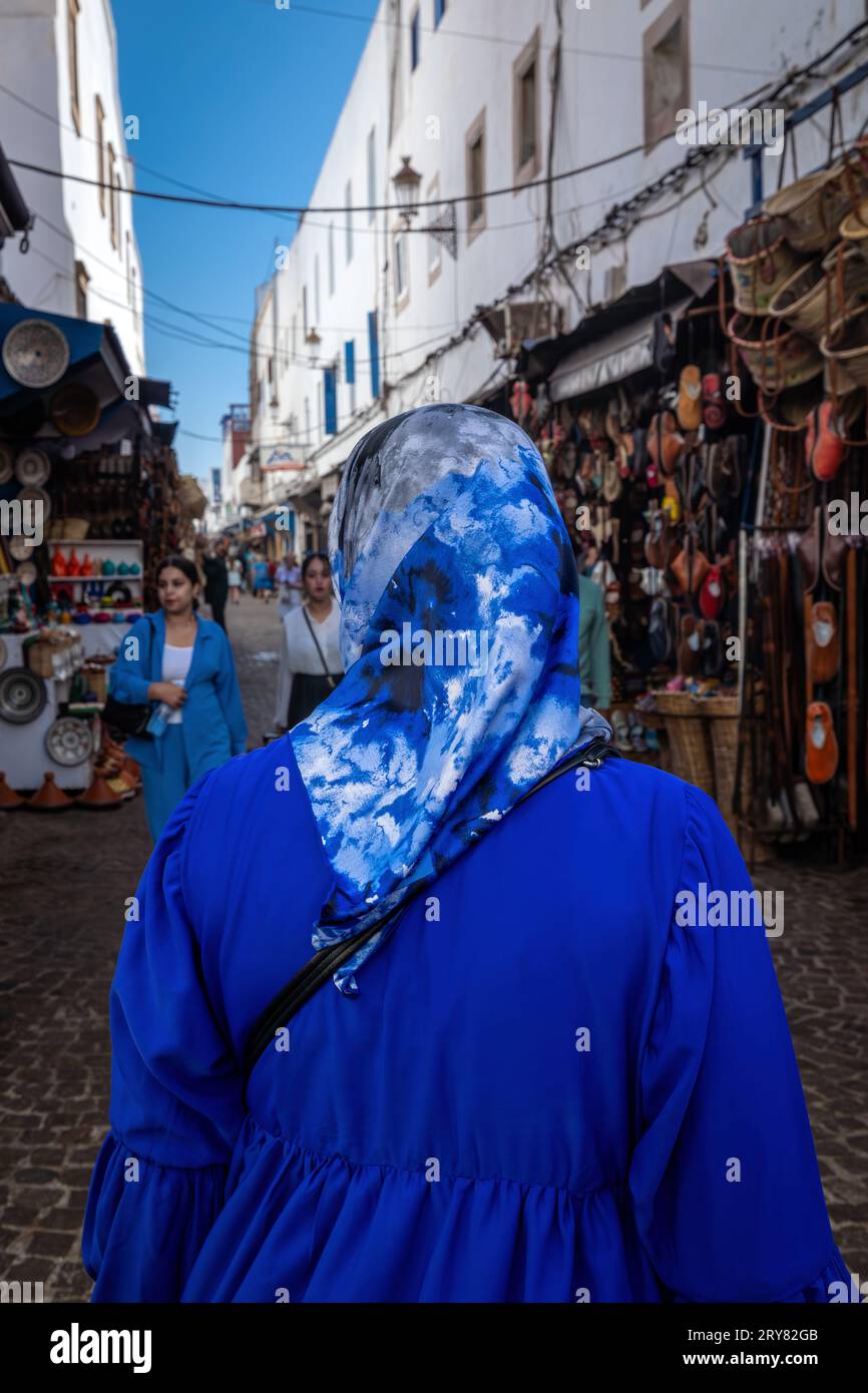 Essaouira, Marocco - 3 agosto 2023: In una stretta strada del centro storico Una donna con un velo islamico, hijab, cammina tra i negozi di artigianato Foto Stock
