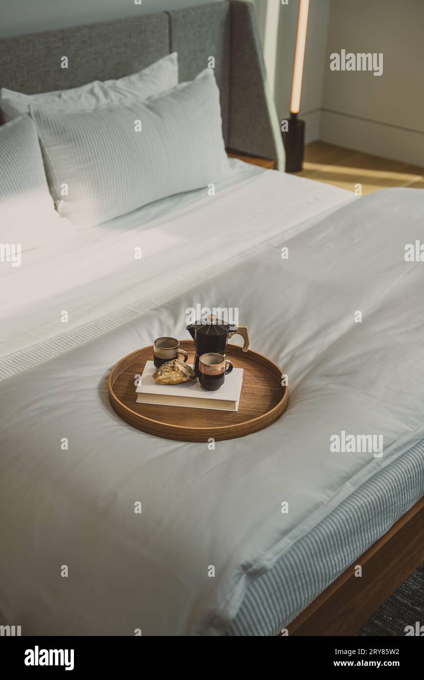 un vassoio da letto su un grande letto king size con biancheria da letto bianca, il vassoio include una caffettiera da 2 tazze di caffè e un libro bianco su un vassoio circolare di legno Foto Stock