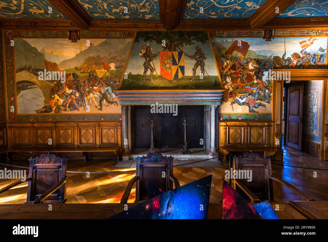 Un camino con uno stemma dipinto affiancato da due cavalieri costituisce il punto centrale di una parete ricoperta da affreschi nella Sala dei Cavalieri (Salle des Chevaliers) del Castello di Gruyères (Château de Gruyères) a Épagny vicino a Gruyères, nel cantone di Friburgo, Svizzera. Scene di battaglia aggiuntive appaiono su entrambi i lati, inclusa una che raffigura combattenti su un ponte. Diversi oggetti angolari a motivi blu e viola di scopo poco chiaro giacciono in primo piano su sedie e sul pavimento. Gli affreschi raccontano la storia leggendaria dei conti di Gruyère dalla loro origine mitica alla battaglia di Morat nel 1476. Il Ginevra Foto Stock