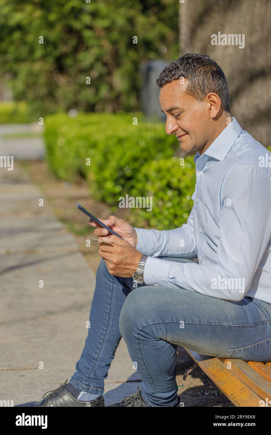 Un giovane latinoamericano che fa un acquisto con una carta di credito sul suo cellulare seduto su una panchina. Foto Stock