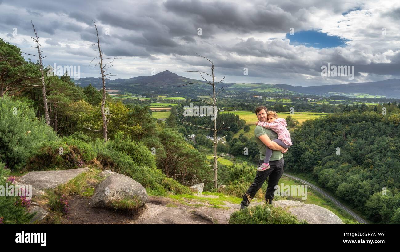 Padre che tiene la figlia in braccio e si trova sul bordo della scogliera, con una vista panoramica su una valle e una pineta. Escursione in famiglia, Wicklow Mountains, Irlanda Foto Stock