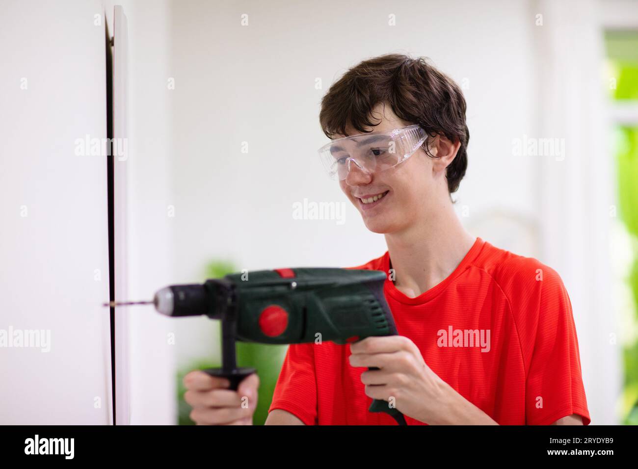 Un giovane che sta perforando una sala in un muro. Ristrutturazione della casa. Appaltatore in funzione. Un ragazzo adolescente che impara a forare. Strumento e utensile per rimodellare. Foto Stock