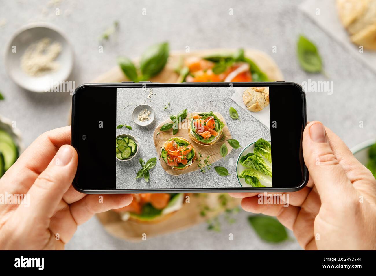 Mani femminili che scattano foto con smartphone di un sano panino con salmone salato, formaggio cremoso, lattuga verde romaina e cetriolo fresco per la pausa Foto Stock