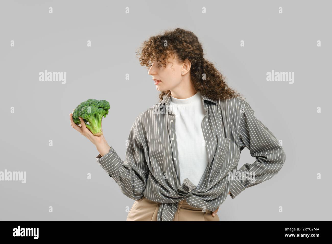 La donna con lunghi capelli ricci tiene i broccoli in mano e li guarda Foto Stock